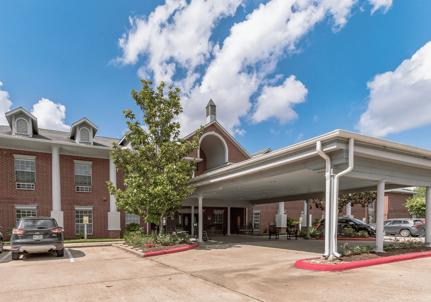 Brick building with a large covered drop-off area and parked cars under a bright blue sky.