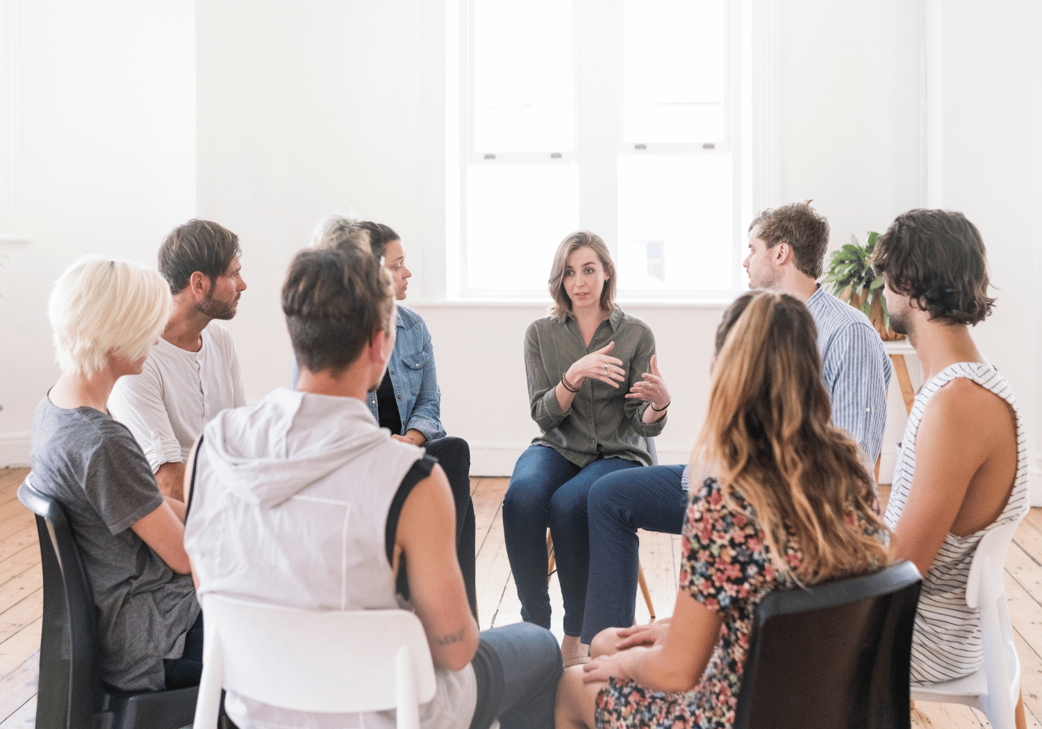 A woman leads a group discussion as eight people sit in a circle in a bright, sunlit room.